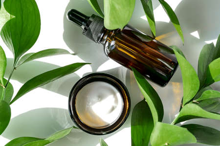 Moisturizer cream jar, serum in amber glass bottle and green leaves on white background. Flat lay,...