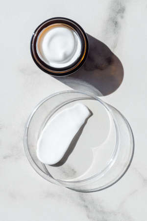 Face cream in a jar and moisturizer smear in petri dish on white marble background. Beauty facial...