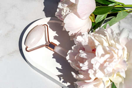 Rose quartz face roller for beauty facial massage therapy. Skin care tool for massage morning routine. Luxury product on white marble background with peony flowers. top viewの写真素材