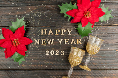 happy new year 2023 inscription made of wooden letter, red poinsettia flowers and champagne glasses on wooden background. Festive greeting card for winter holidaysの写真素材