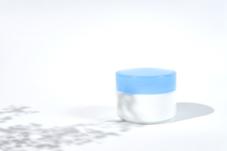 Cosmetic white jar blue lid with cream or lottion and flowers shadows. White background with daylight and harsh shadowsの写真素材