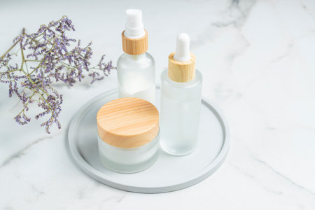 White frosted glass cosmetic bottles and jars with pamboo lids with flowers on ceramic tray on...