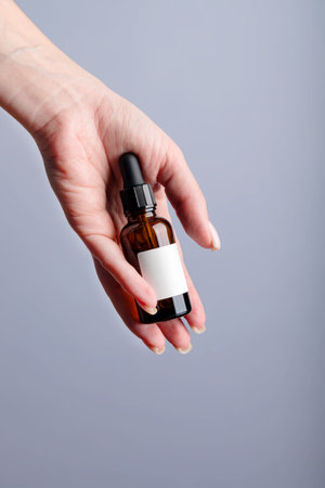 Woman hand holding facial essential oil or serum packaging with blank label on gray background. Beauty cosmetic product for skincare concept. mockup for brandの写真素材