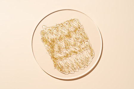 Golden sparkling glitter gel smear in glass petri dish with shadows on beige background for skincare and beauty concepts, laboratory sampleの写真素材