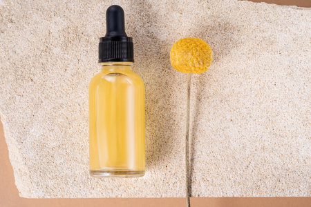 Cosmetic bottle with serum with craspedia flower. Skin care cosmetic. Hyaluronic acid oil, serum with collagen and peptides. unbranded package on sandstoneの写真素材