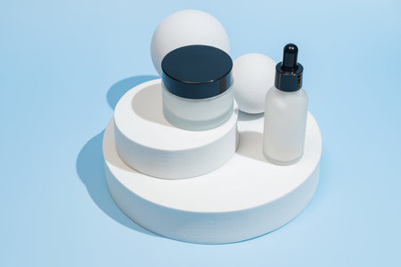 Minimalist white skincare serum dropper bottle and cream jar displayed on a round white podium, showcasing clean beauty packaging. Elegant, luxurious aesthetic perfect for cosmetic brandsの写真素材