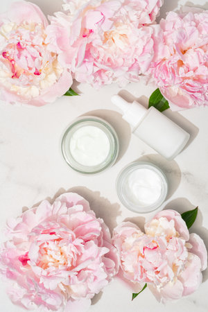 Serum dropper bottle and cream jar with pink peonies, creating a fresh floral skincare flat lay for beauty brands and organic campaignsの写真素材