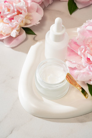 Serum dropper bottle and cream jar on a concrete tray with pink peonies. Urban-meets-natural skincare flat lay for modern beauty brandsの写真素材