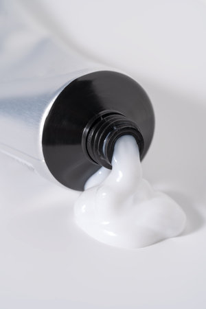 A close-up view of a thick, glossy white cream, gel, or lotion squeezing from a metallic tube onto a...