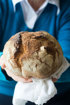 A loaf of bread, homemade. White towel. A woman in blue stand.