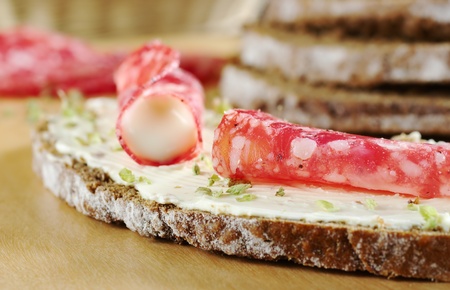 Salami slice and cream cheese with dried herbs on a slice of brown bread on wooden board and ingredients in the background (Selective Focus, Focus on the salami in the front)の写真素材