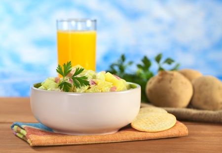 Potato salad made of cooked potatoes, red onions and cucumber, seasoned with a mayonnaise dressing and garnished with a parsley leaf with crackers on the side and orange juice in the back (Selective Focus, Focus on the front of the salad and the leaf)の写真素材