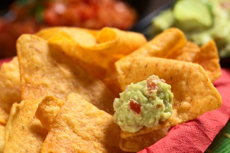 Fresh homemade guacamole (a Mexican sauce made of avocado cream, tomato and onions) on a nacho in a bowl full of nachos (Selective Focus, Focus on the front of the guacamole on the nacho)の写真素材