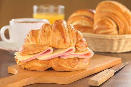 Fresh croissant with ham and cheese on wooden board with coffee, orange juice and bread basket in the back (Selective Focus, Focus on the front of the croissant and the ham and cheese slices)の写真素材