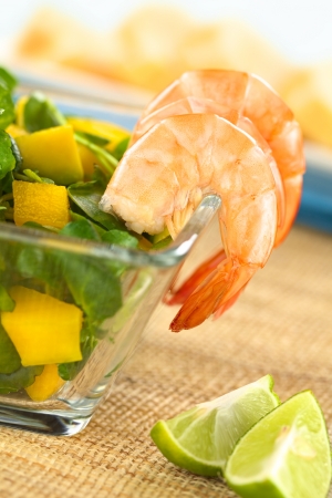 Cooked shrimps on the rim of a glass bowl with fresh watercress, mango and avocado salad with lime wedges on the side and baguette in the back (Selective Focus, Focus on the first shrimp)の写真素材