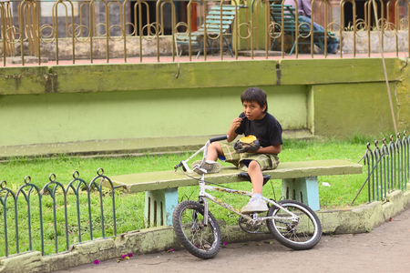 BANOS, ECUADOR - FEBRUARY 22, 2014: Unidentified boy with bike eating a snack while sitting on a bench in Sebastian Acosta Park on February 22, 2014 in Banos, Ecuador. のeditorial素材