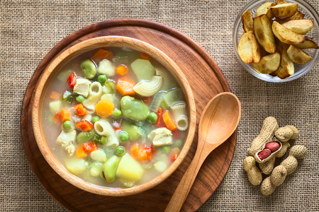 Overhead shot of Bolivian traditional Sopa de Mani (peanut soup) made of meat, pasta, vegetables (pea, carrot, potato, broad bean, pepper, corn) with ground peanut in wooden bowl, traditionally served with fried potatoes, photographed with natural light Overhead shot of Bolivian traditional Sopa de Mani (peanut soup) made of meat, pasta, vegetables (pea, carrot, potato, broad bean, pepper, corn) with ground peanut in wooden bowl, traditionally served with fried potatoes, photographed with natural light