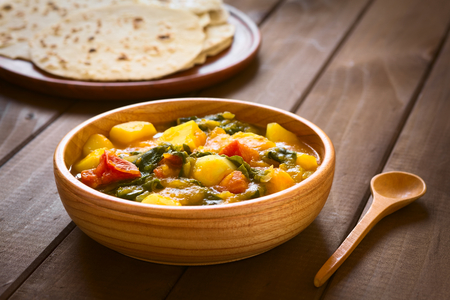 Wooden bowl of pumpkin, mangold, potato and tomato curry dish with homemade chapati flatbread in the back photographed with natural light (Selective Focus, Focus in the middle of the curry dish)の写真素材