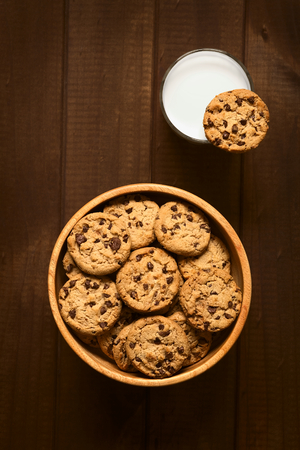 Overhead shot of chocolate chip cookies in wooden bowl and a cookie on the rim of a glass of cold milk, photographed on wood with natural lightの写真素材