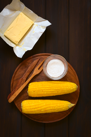 Overhead shot of two cobs of cooked sweet corn on wooden plate with salt and cutlery, with a piece of butter above, photographed on dark wood with natural lightの写真素材