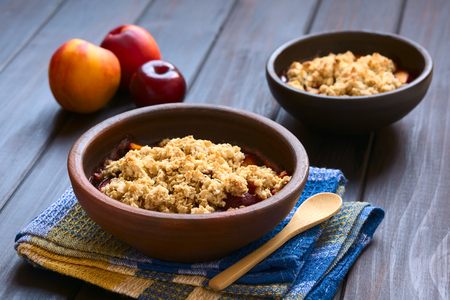 Two rustic bowls filled with baked plum and nectarine crumble or crisp, photographed on dark wood with natural light (Selective Focus, Focus one third into the first dessert)の写真素材