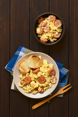 Overhead shot of scrambled eggs made with chorizo slices and onion on plate with toasted baguette slices, wooden fork on the side, photographed on dark wood with natural lightの写真素材