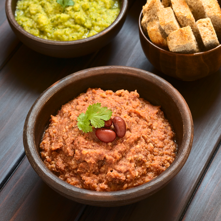 Rustic bowl of homemade red kidney bean spread garnished with kidney beans and fresh coriander leaf, zucchini-parsley spread and sticks of wholegrain bread in the back, photographed with natural light (Selective Focus, Focus on the coriander leaf)の写真素材
