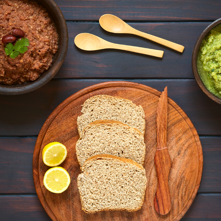 Overhead shot of wholegrain bread slices on wooden plate with two rustic bowls of homemade vegetable spreads (red kidney bean, zucchini and parsley), photographed on dark wood with natural lightの写真素材