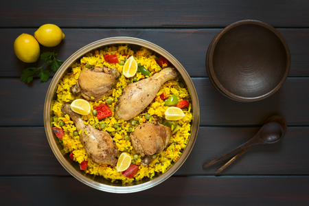 Overhead shot of a pot of chicken paella, a traditional Valencian (Spanish) rice dish made of rice, chicken, peas and capsicum and served with lemon, photographed on dark wood with natural lightの写真素材