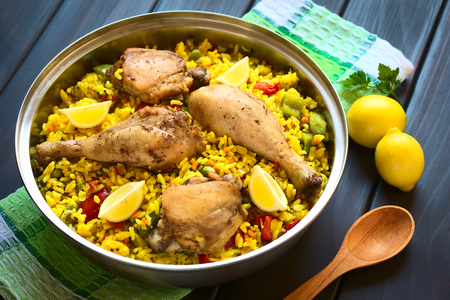 A pot of chicken paella, a traditional Valencian (Spanish) rice dish made of rice, chicken, peas, capsicum and served with lemon, photographed on dark wood with natural light (Selective Focus, Focus on the middle of the dish)の写真素材