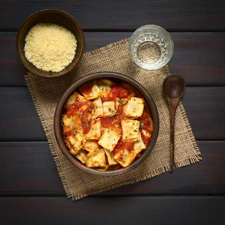 Baked ravioli with homemade tomato sauce in rustic bowl with grated cheese in small bowl, glass of water and wooden spoon on the side, photographed overhead on dark wood with natural lightの写真素材