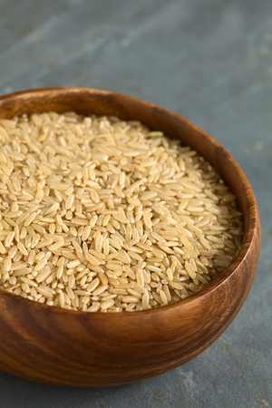 Raw brown or wholegrain rice kernels in wooden bowl, photographed on slate with natural light (Selective Focus, Focus one third into the rice)の写真素材