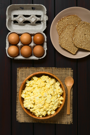 Fresh homemade egg salad prepared with mayonnaise and mustard in wooden bowl, with egg carton and slices of wholegrain bread above, photographed overhead on dark wood with natural lightの写真素材