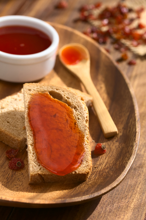 Homemade rose hip jam on wholegrain bread slice on wooden plate with spoon on the side, photographed on dark wood with natural light (Selective Focus, Focus in the middle of the bread slice)の写真素材