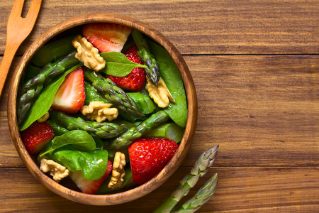 Fresh strawberry, green asparagus, baby spinach and walnut salad served in wooden bowl, photographed overhead on dark wood with natural lightの写真素材