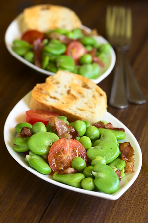 Broad bean, green pea, cherry tomato and fried bacon salad with toasted bread, photographed on dark wood with natural light (Selective Focus, Focus one third into the first salad)の写真素材