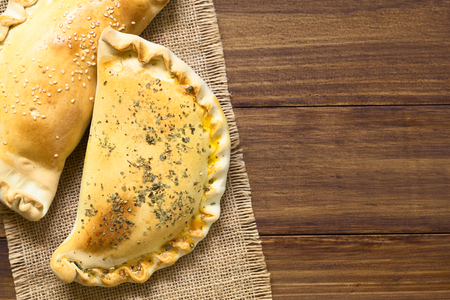 Chilean Empanadas, baked stuffed pastry, photographed overhead on dark wood with natural light. The empanada with the oregano on top is stuffed with ham, cheese, tomato and oregano, the one with the sesame is stuffed with ham and cheese.の写真素材