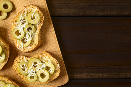 Crostini with green olive slices and freshly grated parmesan-like hard cheese on wooden board, photographed overhead on dark wood with natural light (Selective Focus, Focus on the top of the crostini)の写真素材