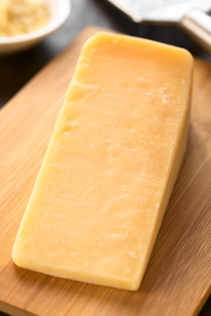 Piece of parmesan-like hard cheese on wooden board with grater in the back, photographed with...