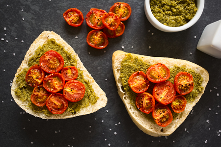 Olive bread roll halves spread with basil pesto and topped with roasted cherry tomato halves, photographed overhead on slate with natural light (Selective Focus, Focus on the top of the rolls)の写真素材