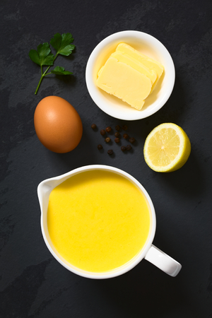 Hollandaise sauce, a basic sauce of the French cuisine, served in a sauce boat with ingredients (egg, butter, lemon, pepper) on the side, photographed overhead on slate with natural lightの写真素材