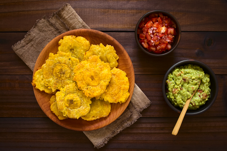 Patacon or toston, fried and flattened pieces of green plantains, a traditional snack or accompaniment in the Caribbean, guacamole and tomato and onion salad on the side, photographed overhead on dark wood with natural light