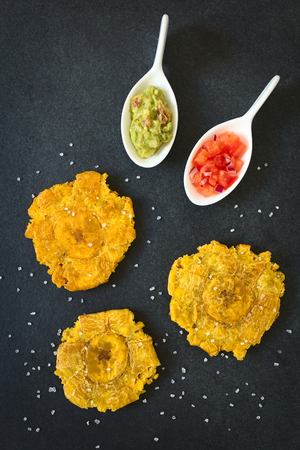 Patacon or toston, fried and flattened pieces of green plantains, a traditional snack or accompaniment in the Caribbean, guacamole, tomato and onion salad on the side, photographed overhead on slate with natural lightの写真素材