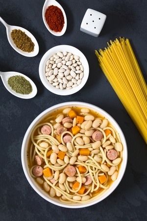 Chilean traditional Porotos con Riendas (beans with reins) dish of cooked dried beans with pumpkin, onion, spaghetti, sausage, with ingredients on the side (cumin, paprika, oregano, white dried beans, spaghetti) on the side, photographed overhead on slate with natural lightの写真素材