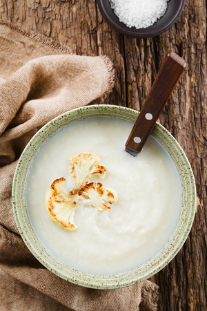 Fresh homemade cream of cauliflower soup garnished with roasted cauliflower floret slices, served in bowl with spoon, photographed overhead on rustic wood (Selective Focus, Focus on the soup)の写真素材