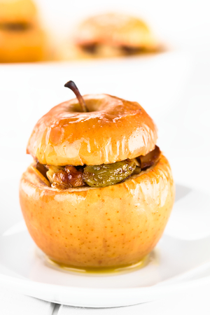 Fresh baked stuffed apple with walnut, almond, raisin, sultana, butter, sugar and cinnamon, a traditional autumn and winter dessert, photographed on white wood (Selective Focus, Focus on the front of the apple)の写真素材
