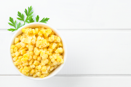 Frozen sweet corn kernels in white bowl, parsley leaves on the side, photographed overhead on white wood with copy space on the right side (Selective Focus, Focus on the corn kernels)の写真素材