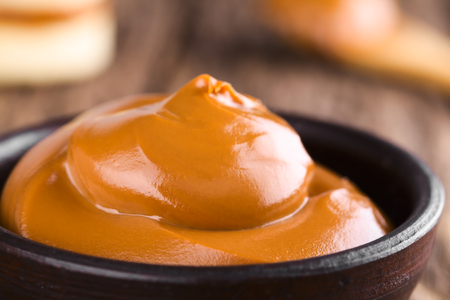 Latin American sweet caramel-like Manjar or Dulce de leche used as spread or filling in baking, photographed in small bowl with alfajor cookie and a spoonful of manjar in the back (Selective Focus, Focus on the front of the cream)の写真素材