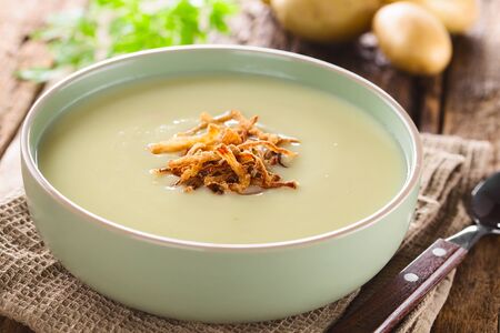 Fresh homemade cream of potato soup in bowl garnished with crispy onion strings, potatoes in the back (Selective Focus, Focus on the onion strings in the front)の写真素材