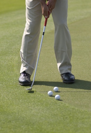 A golfer wearing beige pants and black leather shoes practicing his putt with several balls on a...